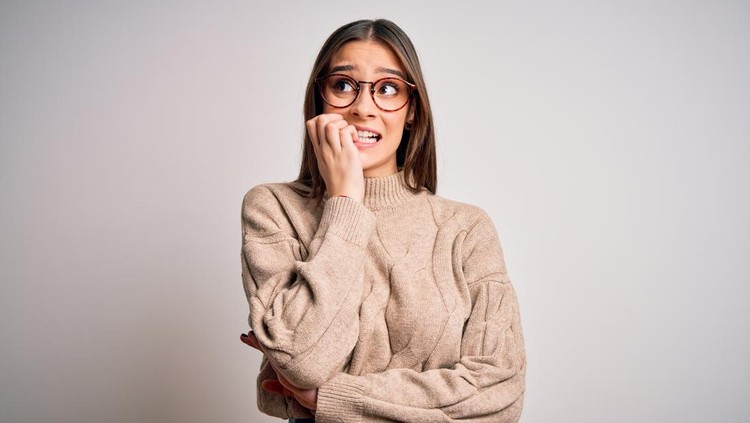 Young beautiful brunette businesswoman wearing casual sweater and glasses standing looking stressed and nervous with hands on mouth biting nails. Anxiety problem.