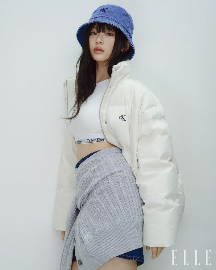 Hanni tampil dengan gaya kasual khas Calvin Klein Jeans. Ia mengenakan jaket puffer cropped putih dan melilitkan kardigan rajut cropped di pinggangnya, memberikan kesan detail yang menarik dari outfit-nya. / Foto: Elle Korea