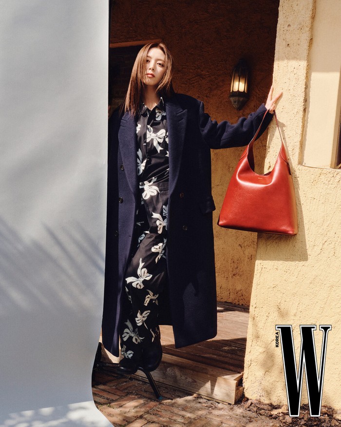 Yuna tampil efforless chic dengan mantel panjang navy yang tebal, dipadukan dengan outfit bermotif pita putih yang menambah kesan feminin. Ia membawa tas kulit besar berwarna merah bata yang memberi sentuhan kontras pada tampilannya./ Foto: W Korea