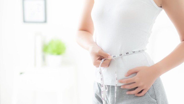 Beautiful young woman measuring her waist with a measuring tape in the living room at home.