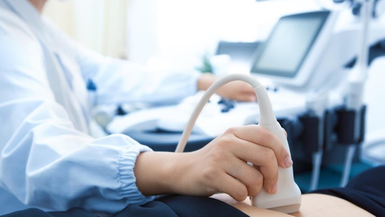 A photo with the focus on the unrecognizable pregnant woman in the foreground as the unrecognizable doctor shows her an ultrasound on a digital tablet in the background.