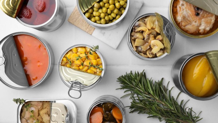 Open tin cans with different products and rosemary on white tiled table, flat lay