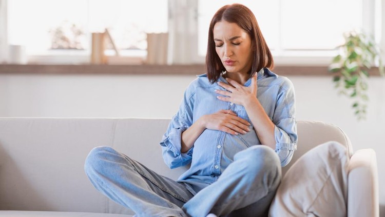 Pregnant lady feeling bad having discomfort and touching her tummy and chest, coping with abdominal spasms and asthmatic issues in living room at home. Complexities of expectant motherhood