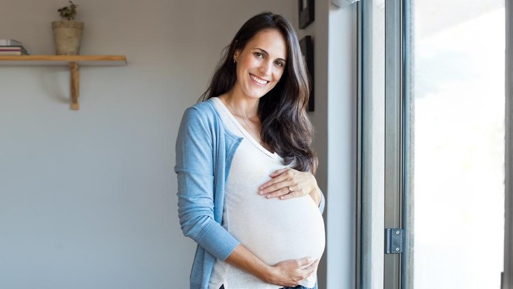 Happy pregnant woman with big tummy relax at home&period; Portrait of pregnant mature woman standing near the window and caresses her belly&period; Smiling woman expecting child and looking at camera&period;