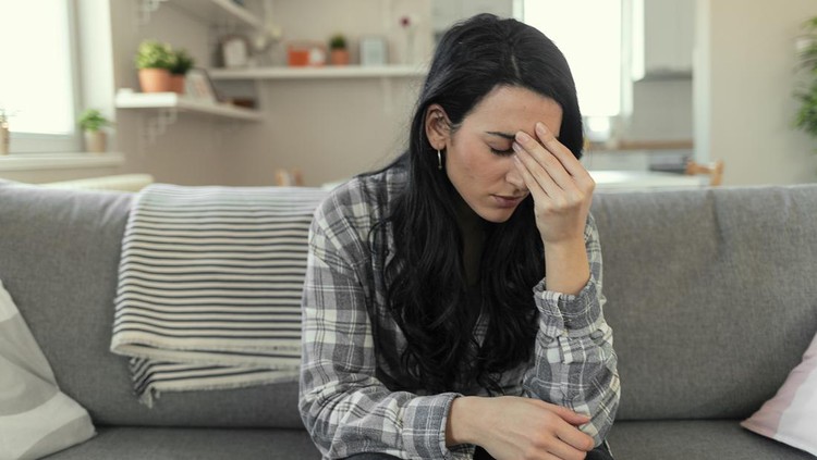 Frustrated young woman suffering from the headache while sitting on the sofa at home with an expression of being unwell, with eyes closed.