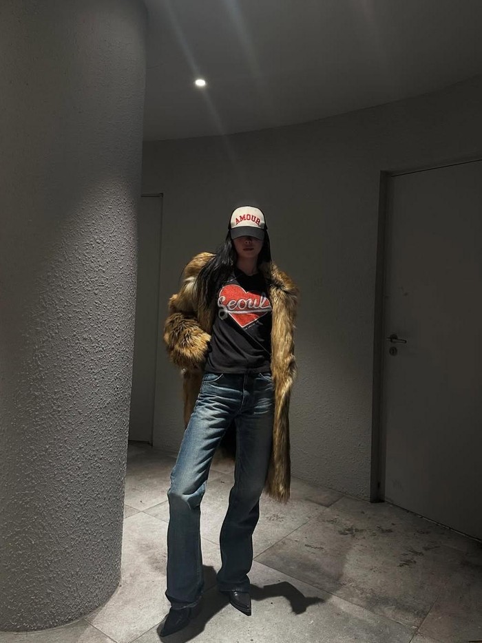 Son Na Eun mengenakan jaket bulu krem yang terlihat sangat hangat dan mewah, memberikan kesan yang glamor. Gabungan jaket bulu, kaus logo, celana panjang, dan topi baseball menciptakan tampilan yang kasual namun tetap stylish./ Foto: Instagram/marcellasne