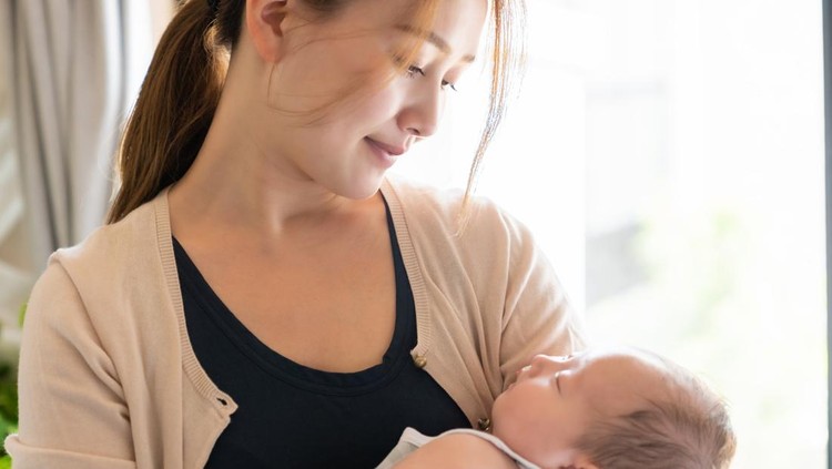 A mother sits in an armchair, holding her newborn in her arms, gazing lovingly at her baby.