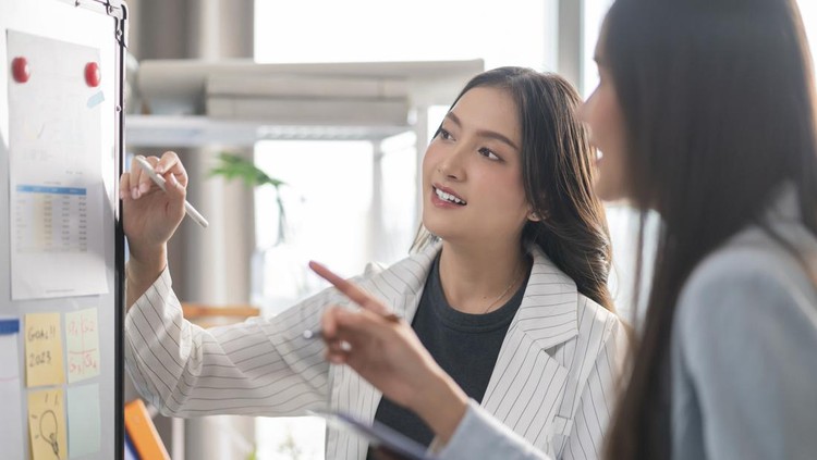 two Businesswoman brainstorming ideas on whiteboard with colleague. Female business partners having brainstorming session about financial budget in startup meeting room.