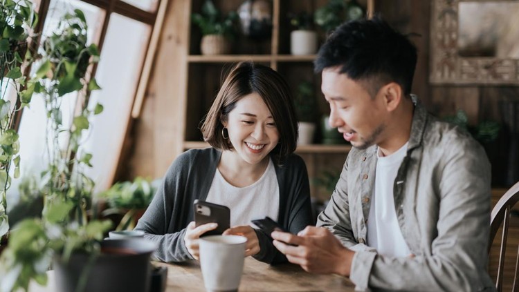 Happy young Asian couple having a coffee date in cafe. Drinking coffee and chatting while using smartphone together. Enjoying a relaxing moment