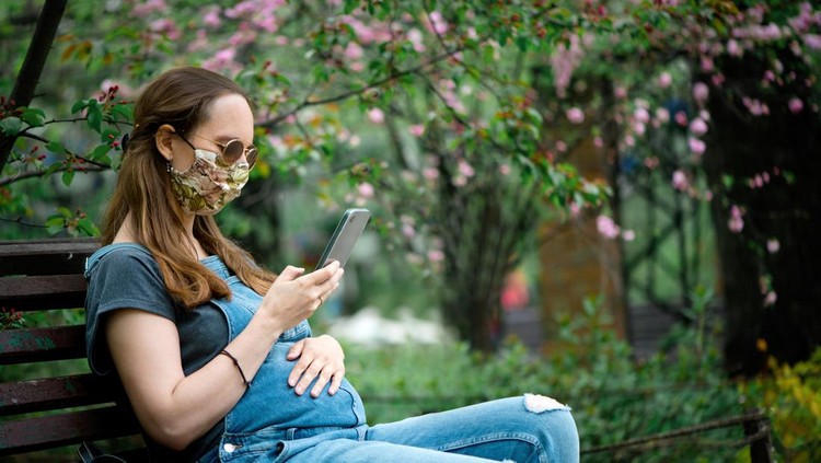 Pregnant woman in protective mask sitting on a bench in springtime in nature.