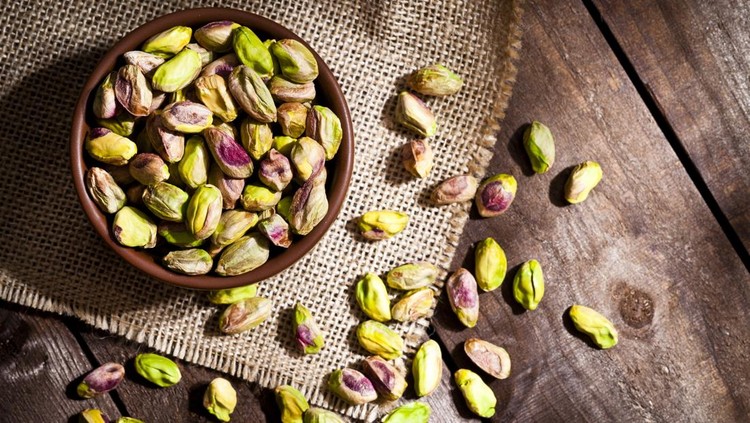Top view of a brown bowl filled with organic pistachios shot on rustic wood table. Some almonds are out of the bowl on a burlap. Predominant colors are green and brown. DSRL studio photo taken with Canon EOS 5D Mk II and Canon EF 100mm f/2.8L Macro IS USM