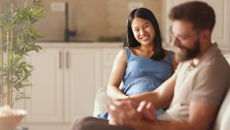 Happy Asian expecting woman enjoying in foot massage by her caring husband on sofa in the living room.