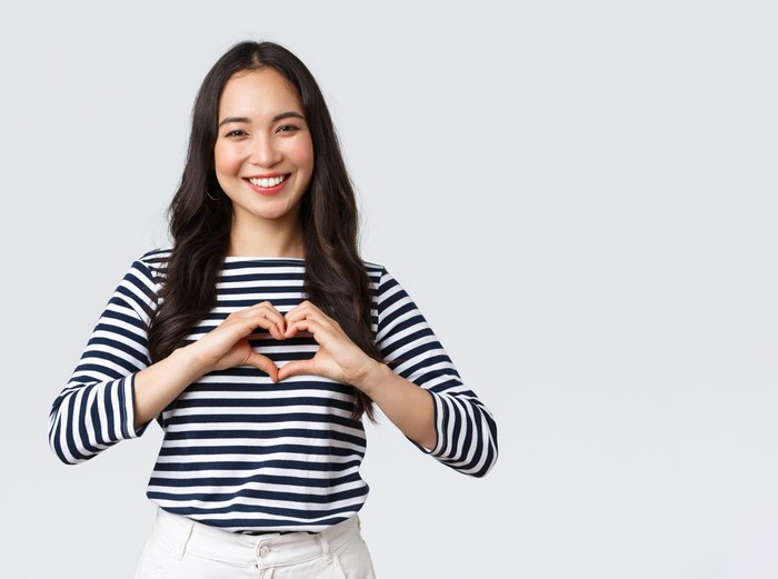 Lifestyle, people emotions and casual concept. Lovely smiling adorable asian woman showing heart sign and smiling, express sympathy or care, standing white background.