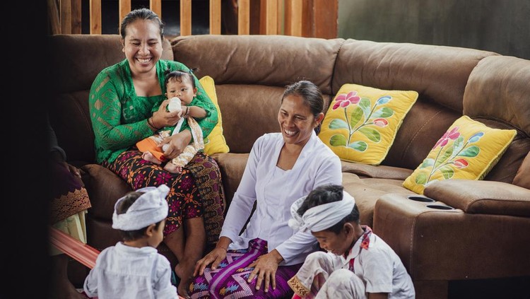 A wide shot of an Indonesian family and their nanny together in the living room at home in Bali, Indonesia. The two youngest boys and the nanny are sitting on the floor while a woman is sitting on the sofa while holding her baby girl. They are all smiling.
