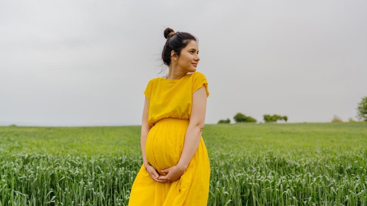 Photo of a pregnant woman standing alone on a grass field, stroking her baby bump and enjoying the calm meadow.