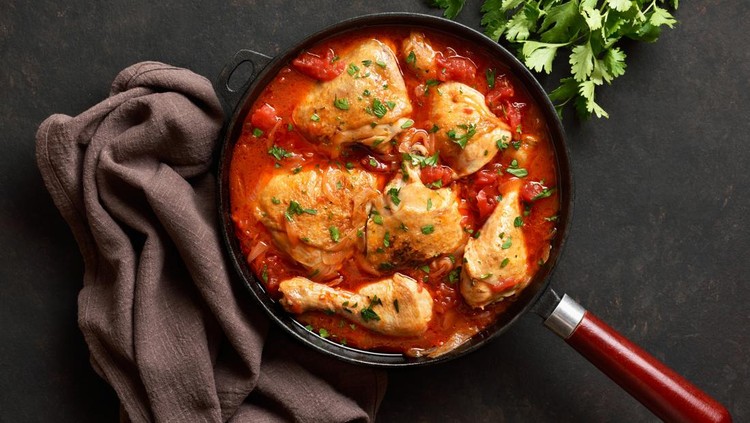Georgian chicken stew with tomatoes and herbs (chakhokhbili) in cast iron skillet over dark stone background. Baked chicken with tomato vegetable gravy. Top view, flat lay