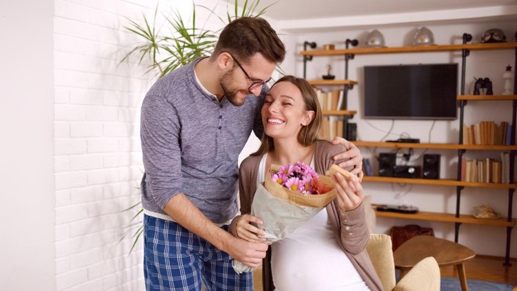 Happy husband surprising his pregnant wife with bouquet of flowers during breakfast