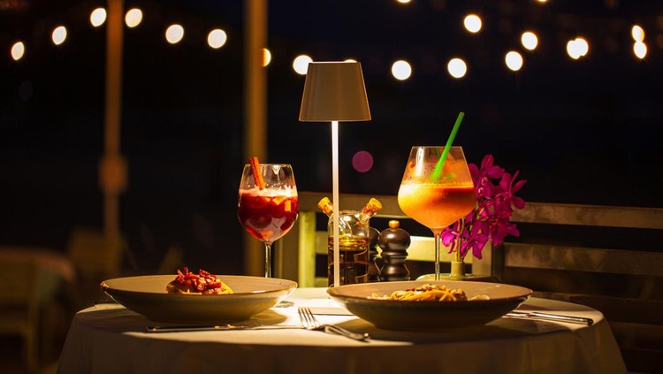 Ilustrasi dinner atau makan malam beachside dinner table,Beachside dinner table with one large challah, two bottles of red wine, two wine glasses, two tea lights in jars, one small vase with flowers, at night, with hanging light nearby in background.