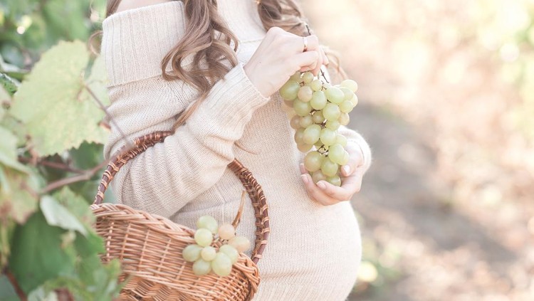 Pregnant woman holding basket and grapes wearing beige knitted sweater outdoors. Harvesting time. Autumn season. 20s.