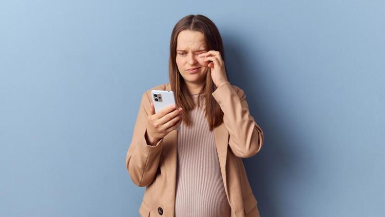 Sad tired young adult pregnant woman wearing dress and jacket posing isolated over blue background rubbing painful eyes after long hours using smartphone grimacing from unpleasant feelings