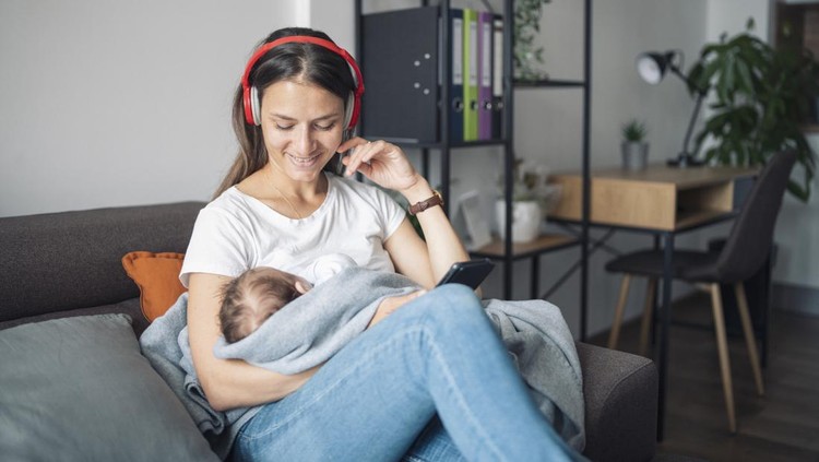 A shot of a young mother using headphones and phone while relaxing and holding her sleeping baby.