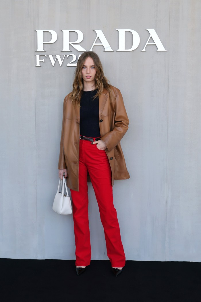 Maya Hawke turut menyaksikan fashion show dalam busana bernuansa maskulin, tapi dengan menggabungkan 4 warna atraktif. Atasan hitam dipadu celana merah yang mencolok. Single-breasted leather coat dan tas putih melengkapi OOTD. / Foto: Courtesy of Prada