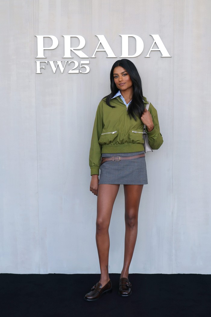 Gaya utilitarian ala Simone Ashley dapat sentuhan chic! Kemeja biru dengan kerah mengintip dari balik cropped jacket hijau yang dilengkapi kantung zipper dikenakannya bersama tailored mini skirt yang punya ilustrasi belt kecil. Penampilannya makin feminin dengan perpaduan loafers dan shoulder bag putih/ Foto: Courtesy of Prada