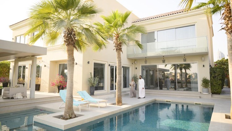 Mid adult man and mature woman wearing traditional Arab clothing on poolside with large house and palm trees