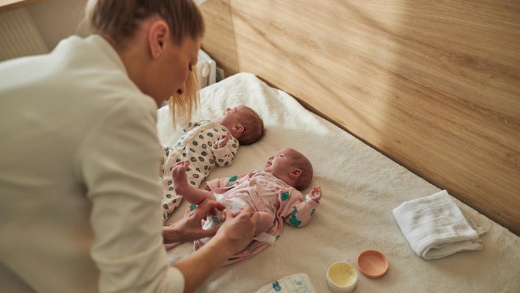 Single mother is taking care of newborn twin babies. She is changing their clothes in bedroom.