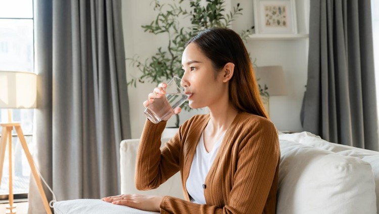 Ilustrasi minum air mineral Happy young Asian woman drinking water while sitting on the sofa in the living room