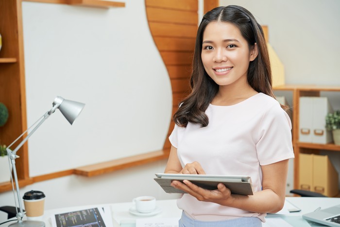 Charming modern Asian woman holding touchpad and smiling at camera in new office