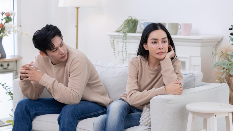 A couple sits on a couch  looking upset. One partner covers their face with their hands, while the other looks away, displaying clear signs of tension and disagreement