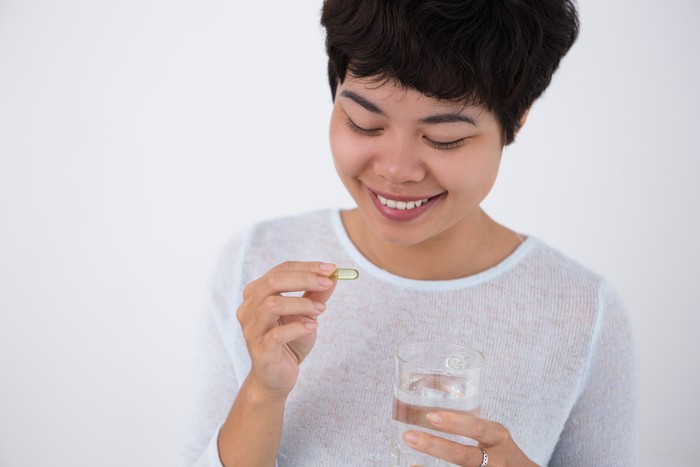 Closeup portrait of smiling young Asian woman holding glass of water and taking pill. Isolated front view on white background.