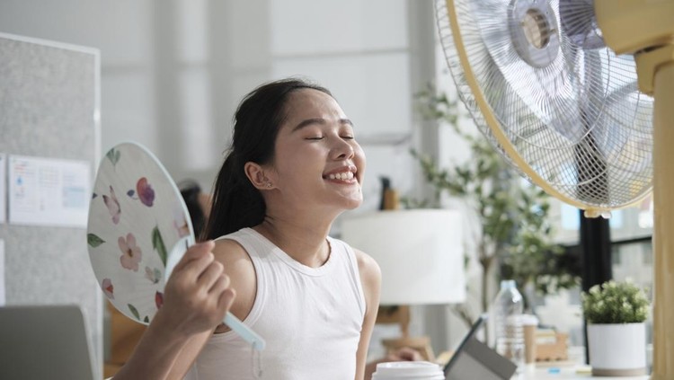 A young female Asian startup worker is blowing wind with electric fan, working in hot temperatures in a casual small business office in summer, refreshment from uncomfortable heat weather season.
