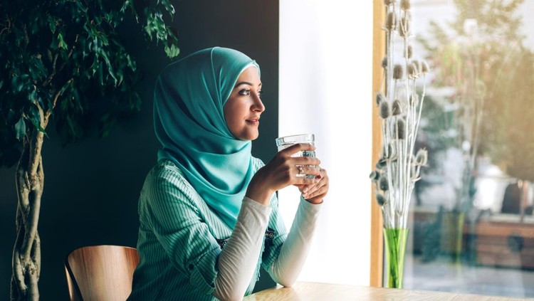 Ilustrasi minum air zam zam Beautiful young arabic girl in hijab posing for camera at cafe with glass of water near the window.