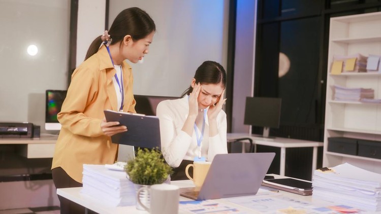 Ilustrasi pebisnis wanita di kantor Two Asian women sit working at desk,overwhelmed with stress struggling with headaches,feeling mentally and physically exhausted while trying to concentrate on their demanding tasks in a busy office