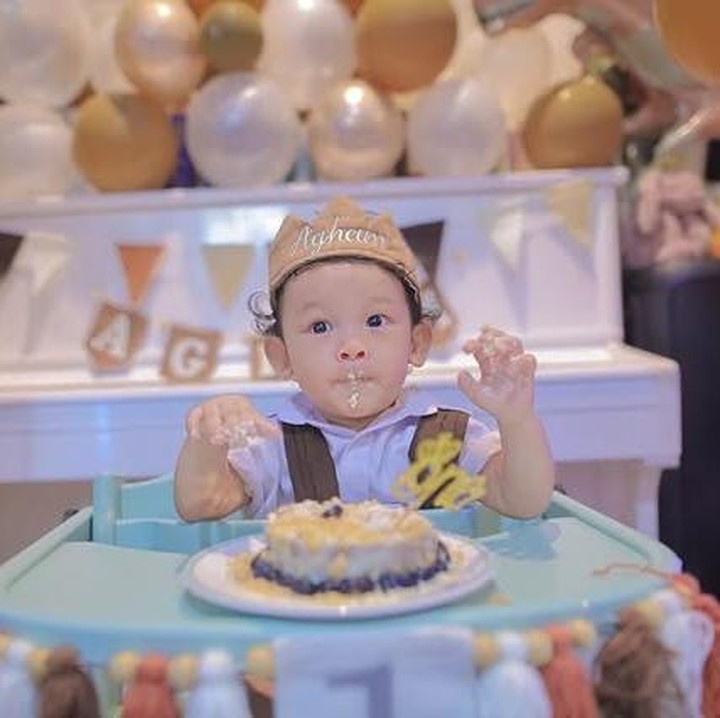 <p>Agham duduk di <em>baby</em> <em>chair</em> dengan ekspresi yang bikin mengemaskan usai mencomot kue ulang tahun pertamanya. Tak hanya itu, Via juga membiarkan putranya mencoba kue ulang tahun tersebut. (Foto: Instagram @viavallen)</p>