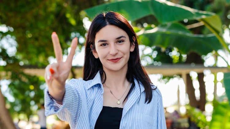 Ilustrasi simbol tangan peace Cheerful brunette woman wearing blue striped shirt on city park, outdoors showing and pointing up with fingers number two while smiling confident and happy. Selective focus on her face.