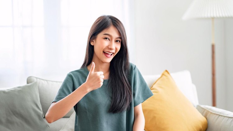 Satisfied asian woman looking at camera with thumbs up sitting on a couch in the living room at home.