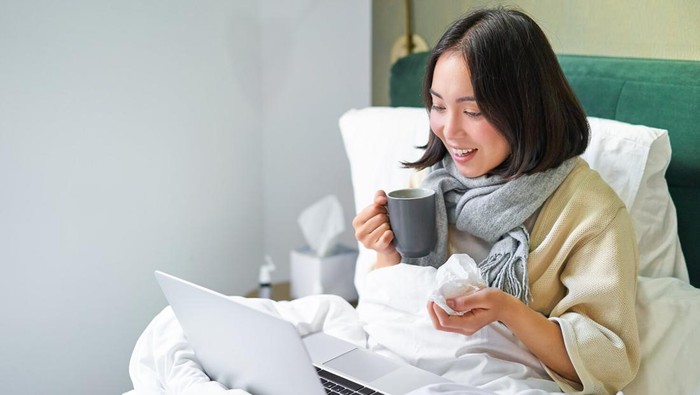 Portrait of asian sick girl, lying in bed with cold, having flu, talking to laptop, video chat, working from home on remote while on sick leave.