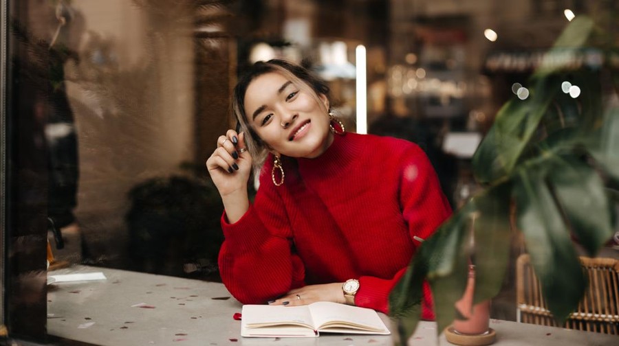 Charming Asian woman in red knit outfit and massive earrings smiles while sitting in cafe with notebook on table