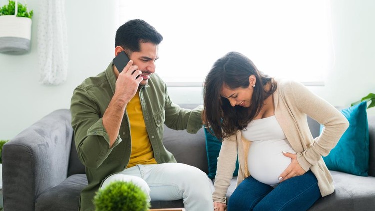 Pained pregnant woman having contractions in the living room and rubbing her belly while her latin husband calls an emergency ambulance on the phone
