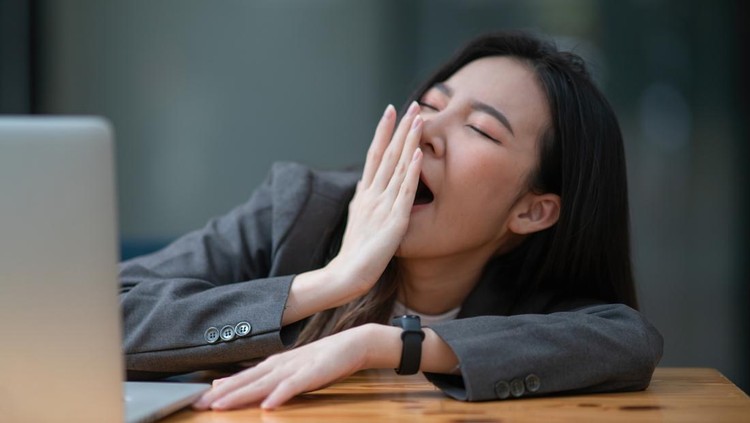 Ilustrasi mengantuk karena sakit diabetes Young businesswoman yawning at a modern office desk in front of a laptop, covering her mouth out of courtesy