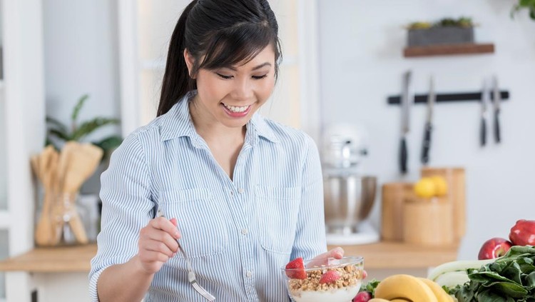 A beautiful young woman looks down and smiles as she stands at her kitchen counter and adds fruit to her yogurt parfait.  There is other fresh produce on the counter.