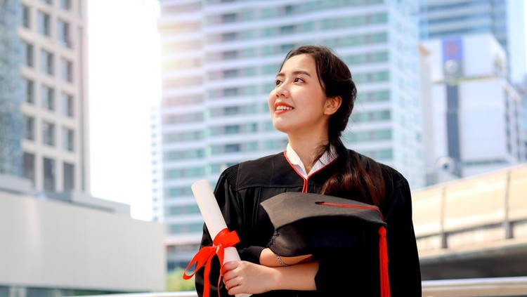 Ilustrasi fresh graduate lulus atau wisuda Happy smiling graduated student, young beautiful Asian woman holding square academic hat cap and certificate, standing with arms crossed at city with skyscraper building background, celebrating successful education on commencement graduation day when finished studies for university or college.