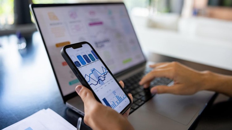 Ilustrasi investasi di pasar saham Close-up on a woman working at home and looking at the stock market behavior on her cell phone and the laptop - business concepts