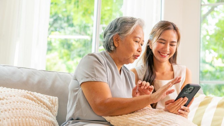 Happy Asian family senior mother and adult child spending time together at home. Daughter teach elderly mom using mobile phone application for online shopping and social media on sofa in living room.