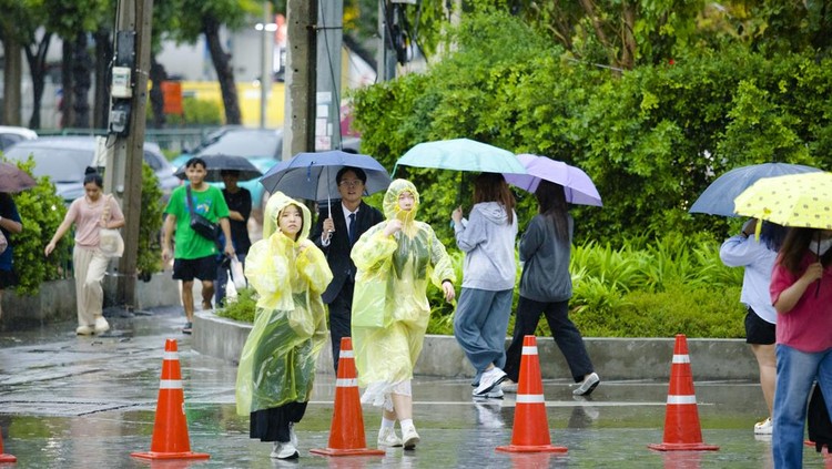 Hujan di bulan Agustus saat kemarauand are on  sidewalk. Some people are using umbrellas. Two young women are walking in yellow raincoats. One woman is cross-eyed. Behind them a man with eywear and in suit is walking.  On street is rush hour traffic