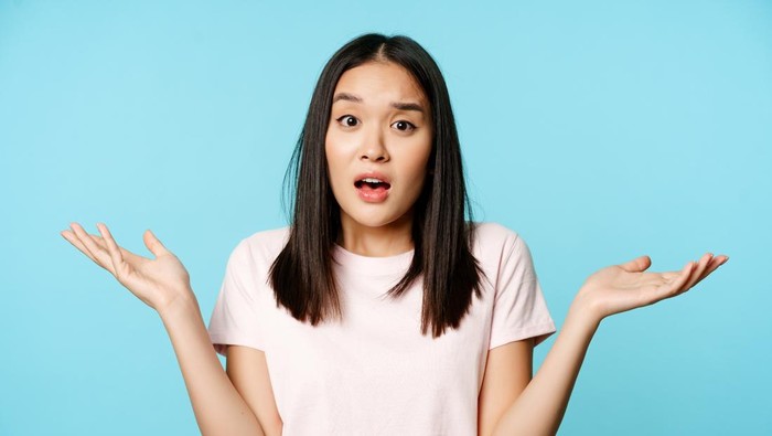 Confused asian girl spread empty hands sideways, shrugging shoulders clueless, standing in tshirt over blue background.