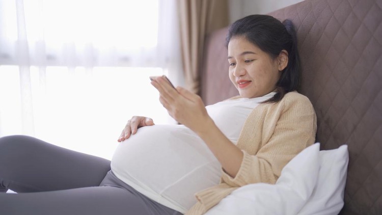 ibu hamil lihat ponsel A portrait of happy Asian pregnant woman using online mobile phone technology device with screen in bedroom at home. Having a baby. Family people lifestyle. Mom love.